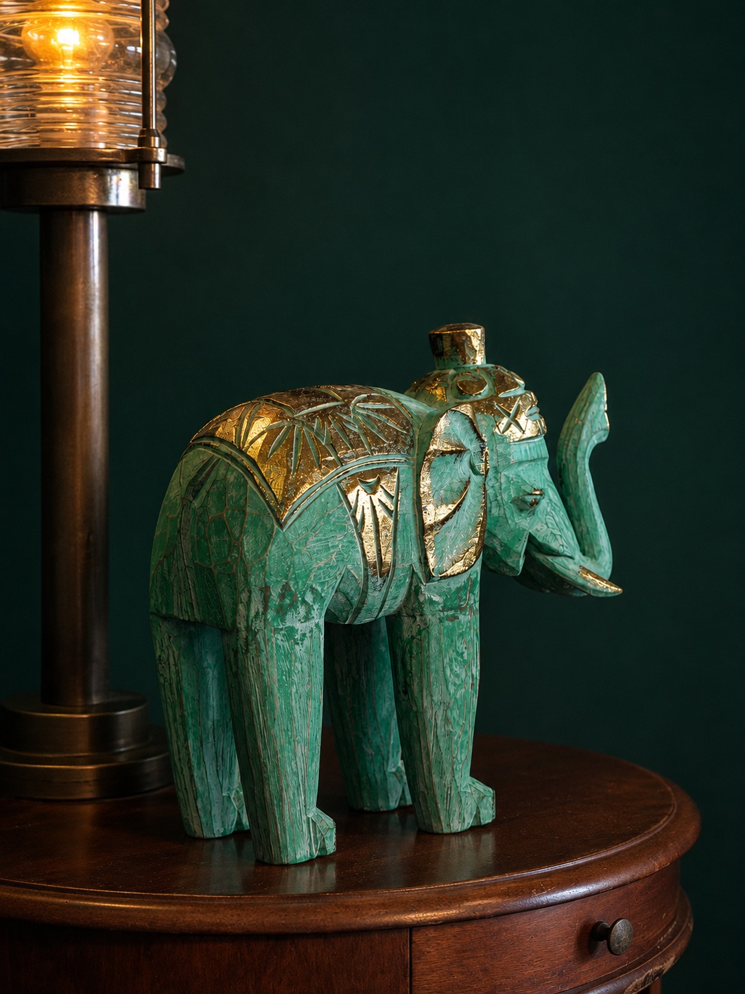 A hand-carved elephant figurine in jade green with gold detailing, standing beside a lit brass lamp on a wooden table
